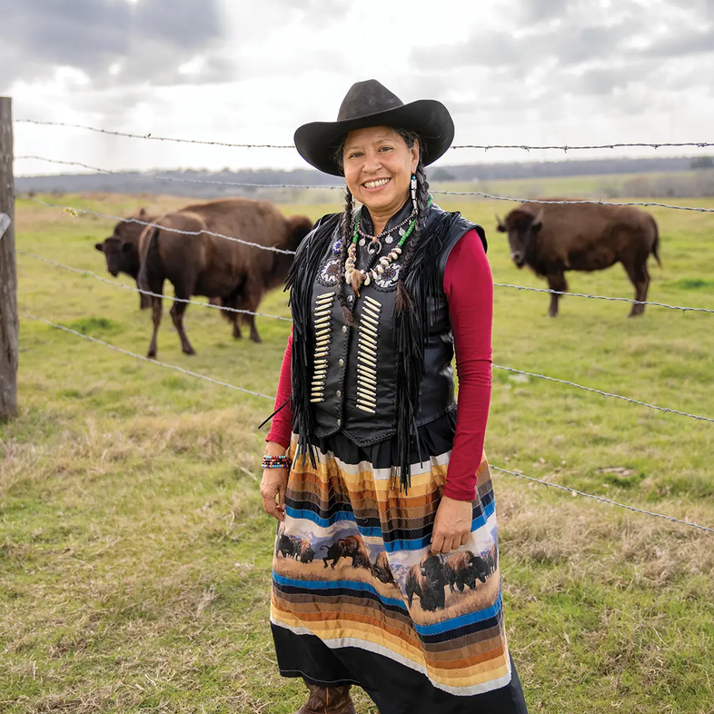 Lucille Contreras dressed in clothing of the Lipan Apache tribe standing in front buffalo in a fence