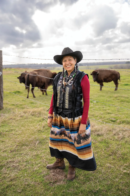 Lucille Contreras dressed in clothing of the Lipan Apache tribe standing in front buffalo in a fence