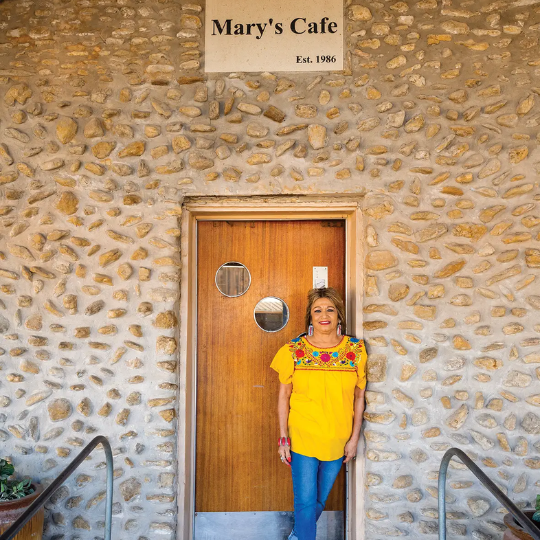 Mary standing at the door of Mary's Cafe at Palo Pinto State Park with a sign reading set. 1986