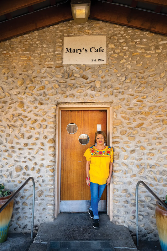 Mary standing at the door of Mary's Cafe at Palo Pinto State Park with a sign reading set. 1986