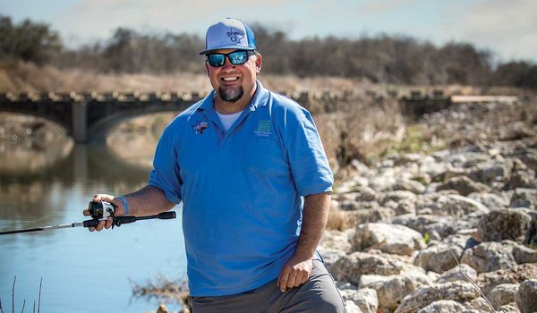 Man in a blue shirt and hat holds a fishing pole as he stands on a rocky river bank