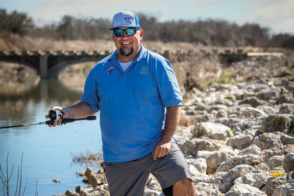 Man in a blue shirt and hat holds a fishing pole as he stands on a rocky river bank