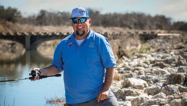 Man in a blue shirt and hat holds a fishing pole as he stands on a rocky river bank