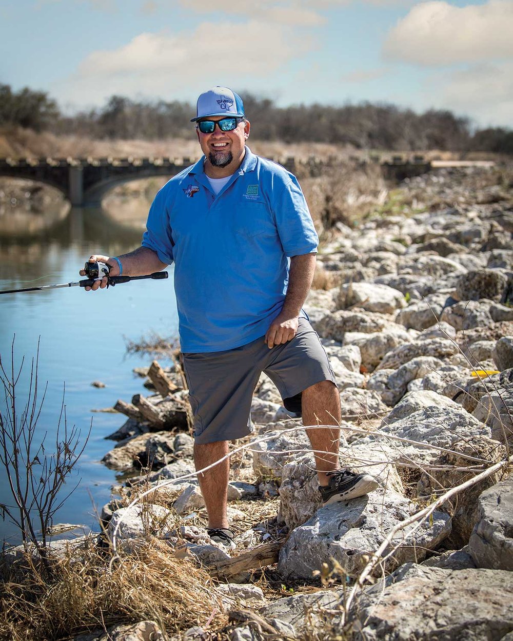 Man in a blue shirt and hat holds a fishing pole as he stands on a rocky river bank