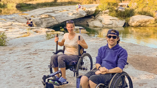 People in all terrain wheelchairs on a rocky trail by water