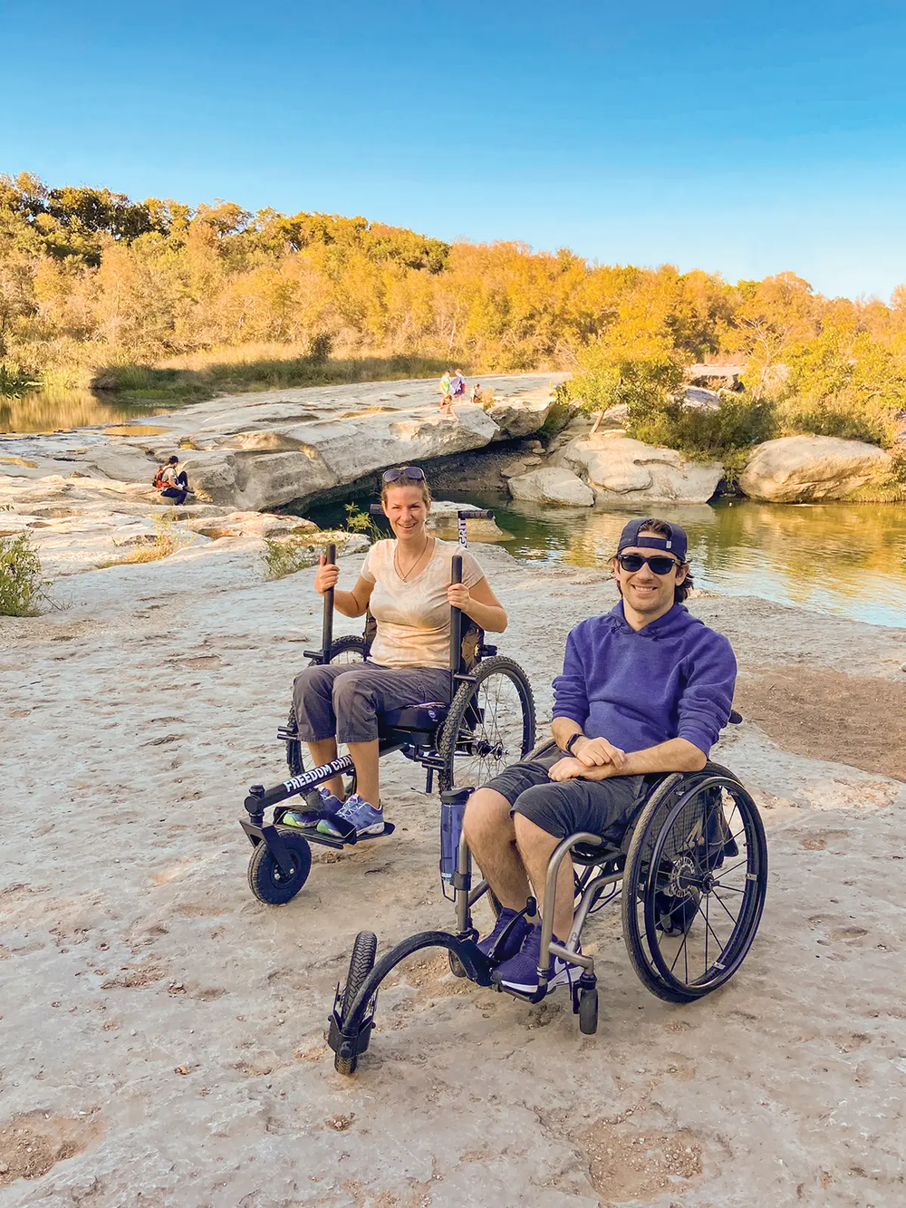 People in all terrain wheelchairs on a rocky trail by water