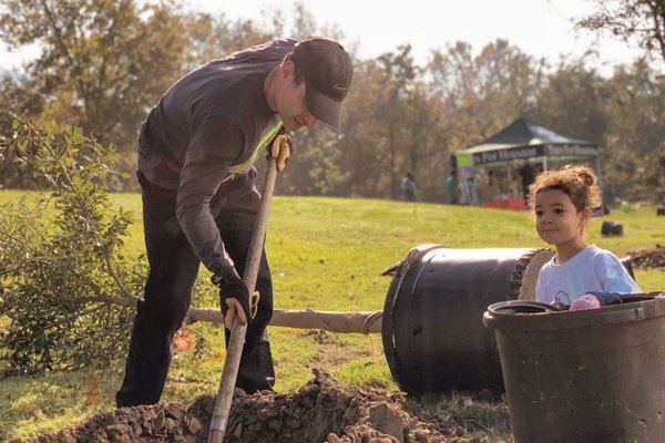 Person digging a hole for a tree and a child standing by the empty pot for the tree.