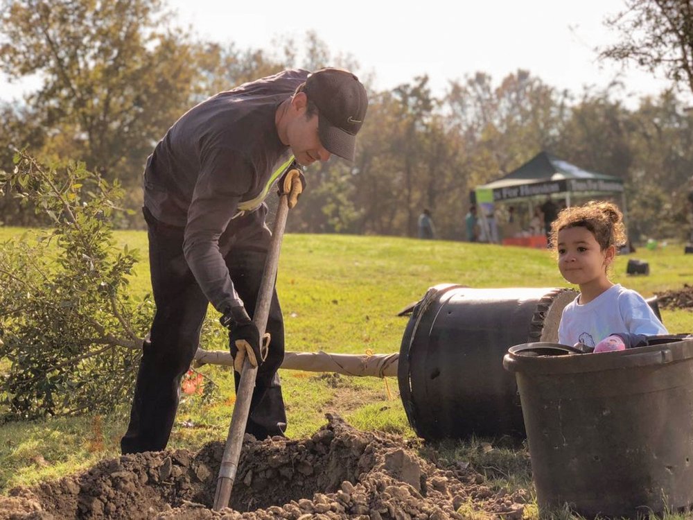 Person digging a hole for a tree and a child standing by the empty pot for the tree.