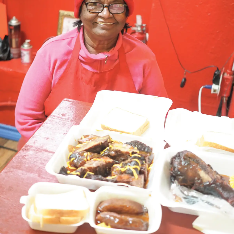 Punarbassi Sandy in Jaws BBQ with a pink hat and shirt sitting at a table with food in togo containers