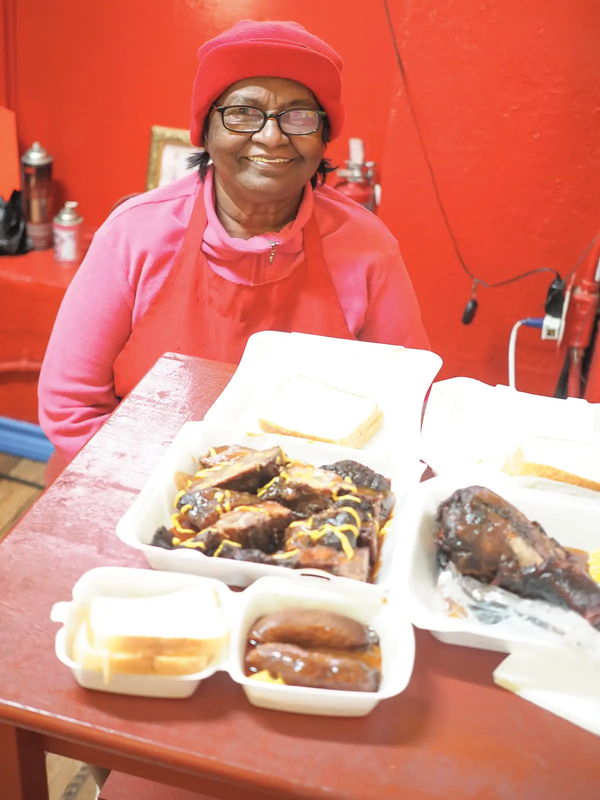 Punarbassi Sandy in Jaws BBQ with a pink hat and shirt sitting at a table with food in togo containers