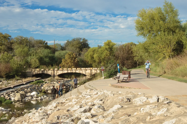 San Antonio River Walk with person riding a bike on the trail while people walk down into tthe rocky channel