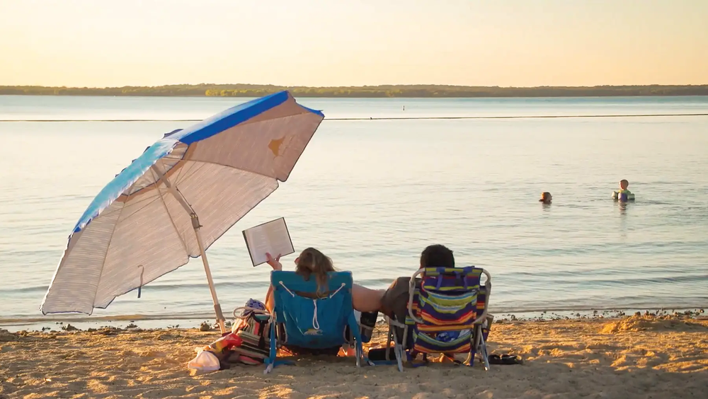 couple on the beach of the lake under an umbrella one reads while the other watches two children swimming