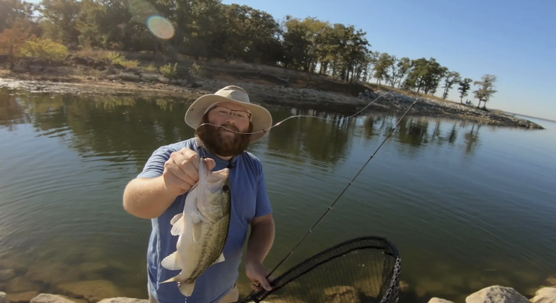 Man in a hat on a rocky river shore holding a net and a fish he caught