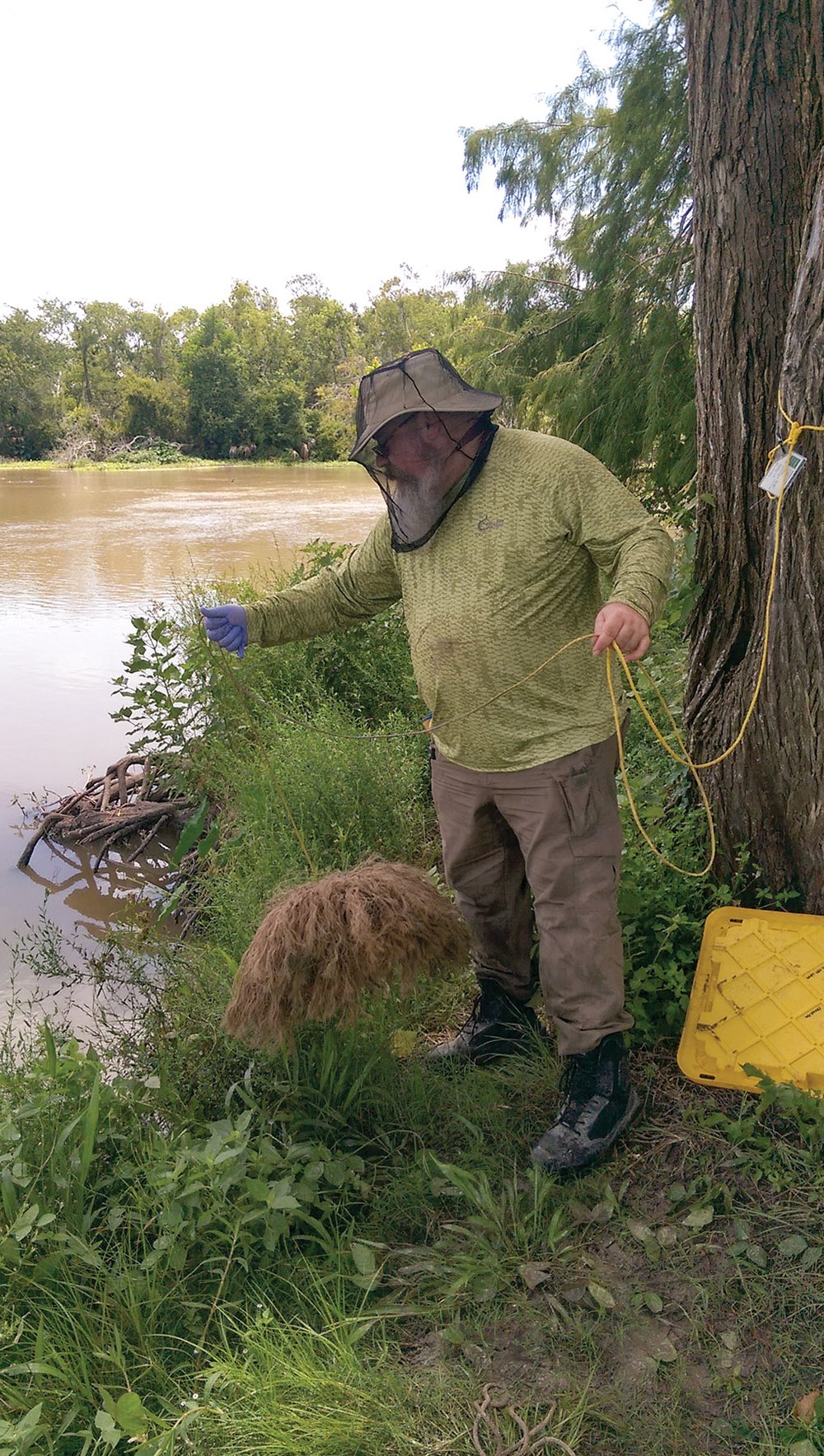 Man in a yellow shirt and khaki pants with a hat and mesh face covering using and eel mop attached to a yellow rope in an attempt to sample the water.