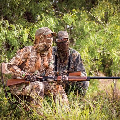 Two people in camouflage kneeling down while hunting turkey.