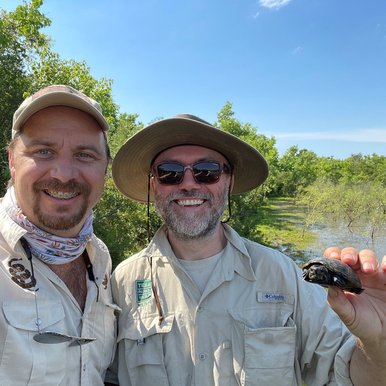 Two herpetologist near water with one holding a tiny turtle.