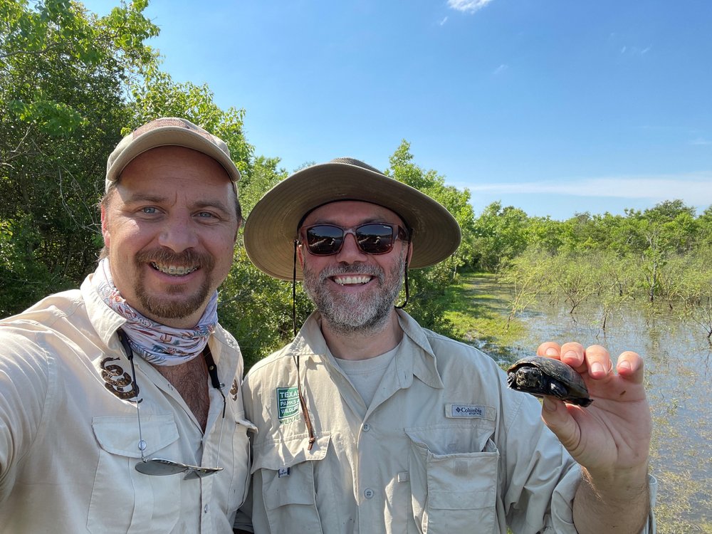 Two herpetologist near water with one holding a tiny turtle.