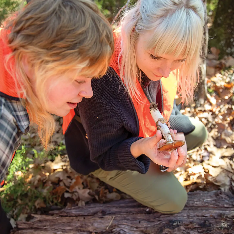 Two women one kneeling on log looking at a long stemmed orange top mushroom