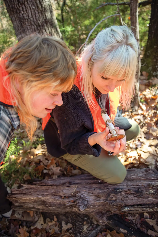 Two women one kneeling on log looking at a long stemmed orange top mushroom