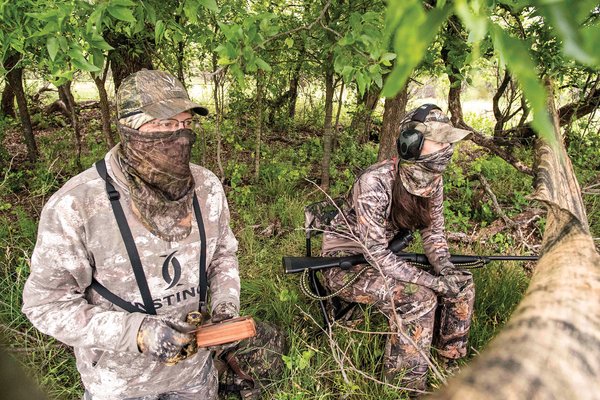 Two hunters behind a tree limb holding a turkey call.