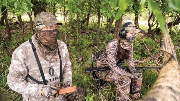 Two hunters behind a tree limb holding a turkey call.