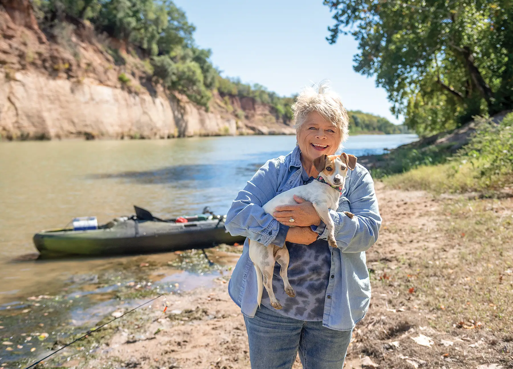 Woman holding her dog with a kayak and the river in the background