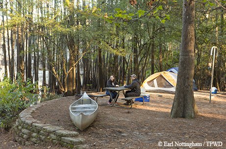 Canoe on a Platue held in place with a stone wall with people eating at a picnic table with a tent pitched in the background in the woods.