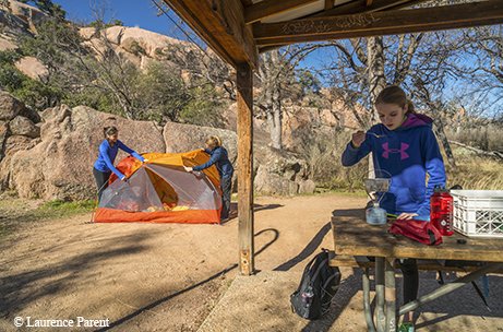 girl standing under a pavilion at a picnic table eating while two people set up a tent with a large rock formation in the background.