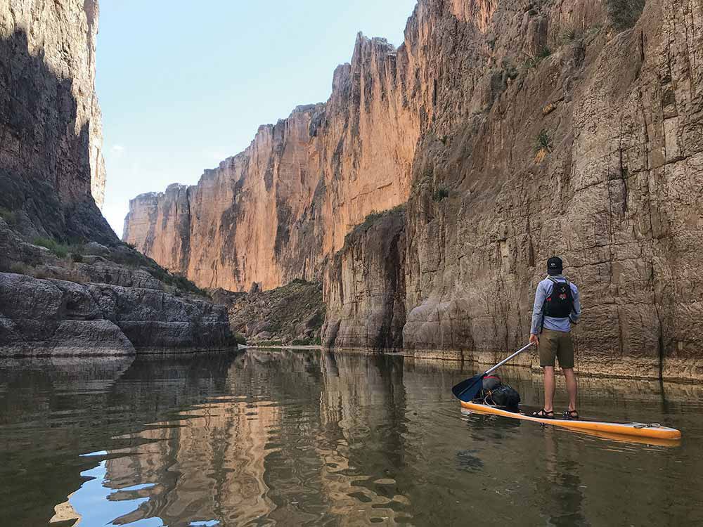 A lone paddle boarder stands on his board, holding an oar, as he makes his way between the cliffs of the Santa Elena Canyon along the Rio Grande