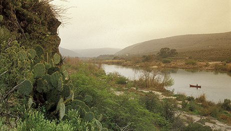 rocky terrain with cacti and with a person in a canoe on the river in the background
