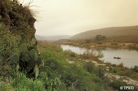 rocky terrain with cacti and with a person in a canoe on the river in the background