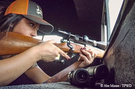 Lindsey Head takes aim at a deer in a blind with binoculars beside her