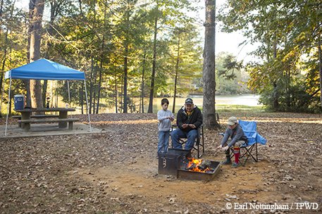Three people around a campfire during the day near a covered picnic table in the woods with a lake in the background