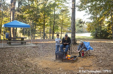 Three people around a campfire during the day near a covered picnic table in the woods with a lake in the background