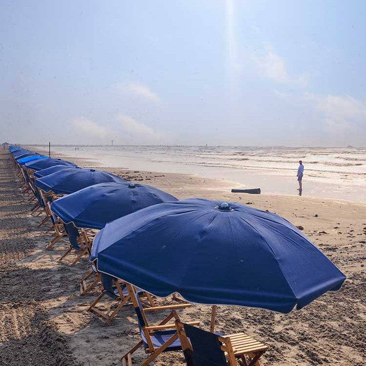 A row of blue beach umbrellas stand over chairs along the sandy shore of the gulf of Mexico