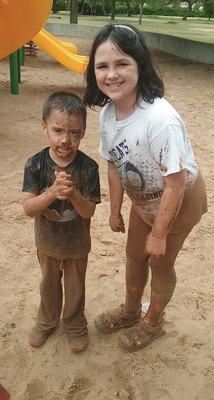 Girl in white playing in the mud with younger sibling who is dressed in black.