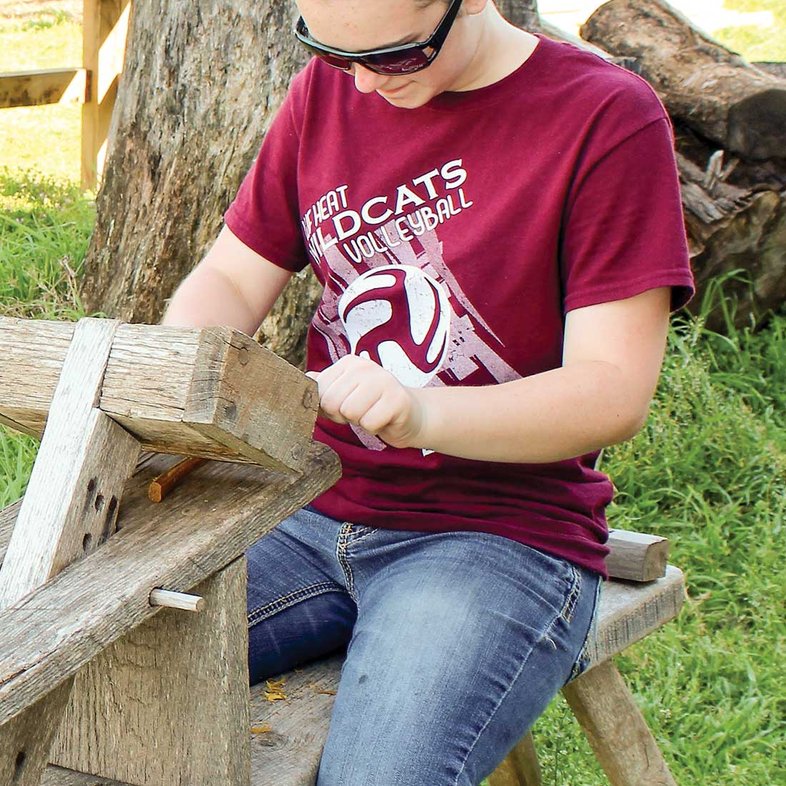 Woman with a red shirt that reads wildcats volleyball sitting on a wooden bench using woodworking tools.