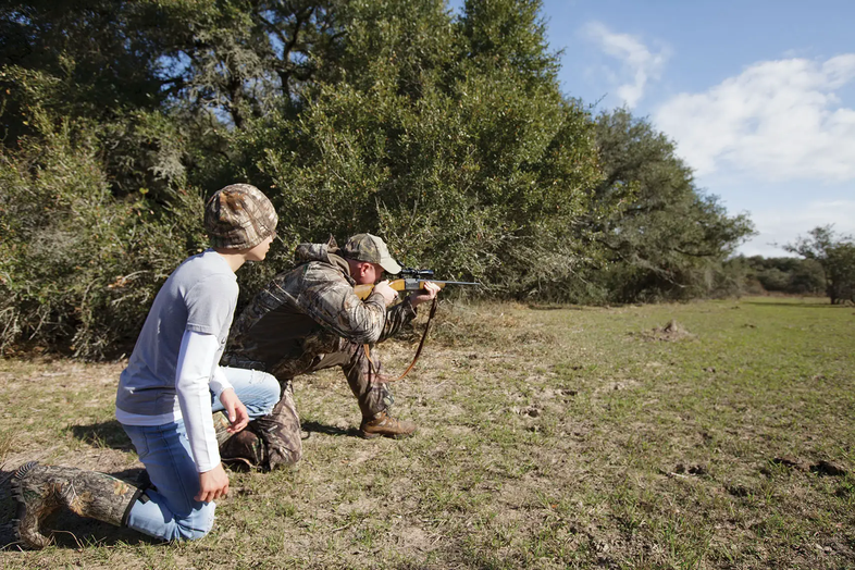 two people kneeling on the ground, one with a gun, while hunting in a field
