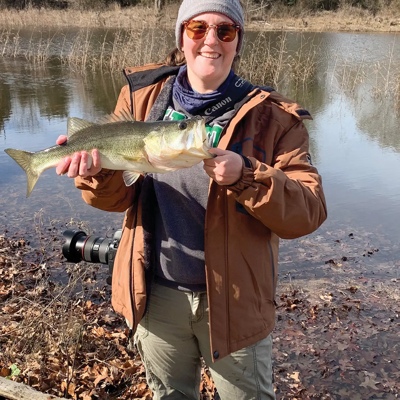 woman with camera holding a bass on the leafy shore of the lake