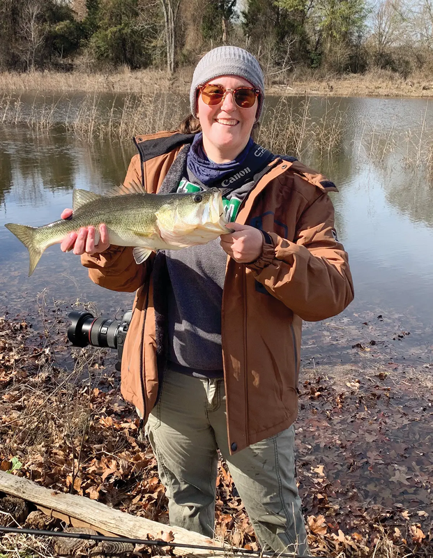 woman with camera holding a bass on the leafy shore of the lake